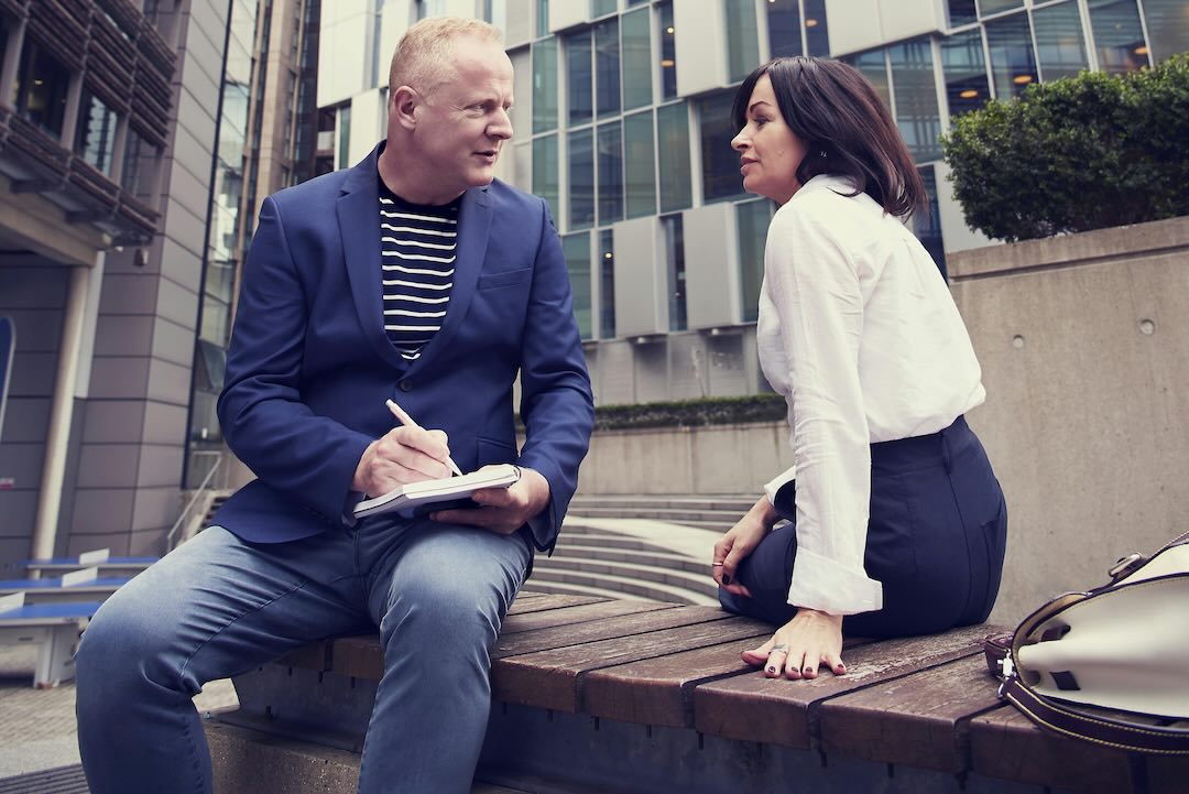 Two professionals having a discussion outdoors on a wooden bench in front of a modern office building; one is taking notes while the other listens attentively.